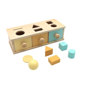 May include: Wooden toy box with three drawers and a lid with cutouts for different shapes. The box comes with a set of wooden shapes, including a circle, square, triangle, rectangle, and a cylinder. The shapes are in shades of yellow and blue.