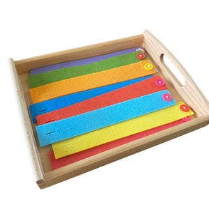 May include: A wooden tray with seven colorful felt strips, each with a button attached. The strips are arranged in a rainbow pattern, with purple, green, orange, blue, red, yellow, and pink.