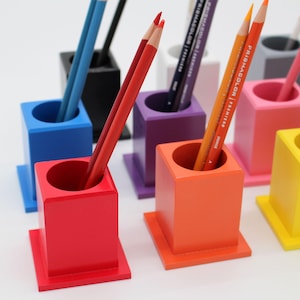 May include: A set of colorful pencil holders in various colors, including red, orange, yellow, pink, purple, blue, and black. The holders are made of wood and have a square base with a round opening for pencils.