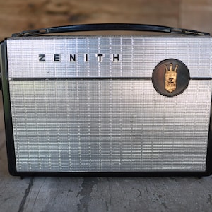 May include: Vintage Zenith radio with a silver mesh front and black trim. The word "ZENITH" is displayed across the top. A circular emblem with a crown and the letter "Z" is on the right side. The radio has a black handle.