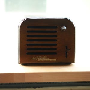 May include: A dark brown wooden speaker with a horizontal slatted front grille, a silver circular knob, and a vertical slider switch. The text "Ampli-fi" and "THE ROLLING TONE" is printed in gold lettering on the lower front of the device.