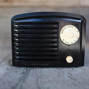 May include: Black vintage Silvertone radio with a cream-colored dial and tuning knob. The radio has horizontal speaker grill lines and the word "SILVERTONE" is printed on the bottom. The dial displays radio frequency numbers.