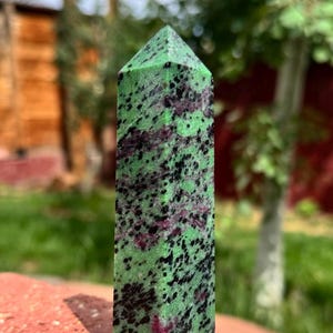 May include: A tall, rectangular zoisite crystal with a pointed top. The stone is a vibrant green with black and maroon speckles. It stands on a red brick surface, with a blurred green background.