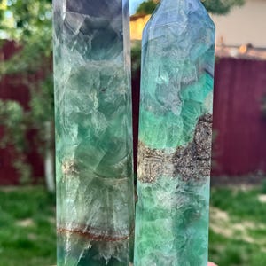 May include: Two tall, polished fluorite crystal towers. The crystals display a range of colors, including green, purple, and clear. The crystals are standing on a brick surface, with a blurred background of greenery and a red fence.