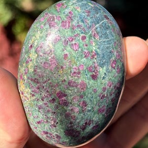 May include: A polished, oval-shaped ruby in zoisite gemstone featuring a mottled green and blue base with embedded, irregular deep magenta and red crystal inclusions throughout the smooth, reflective surface of the stone.
