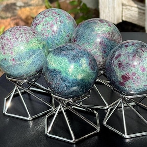 May include: Five polished spheres made of ruby in fuchsite, each featuring a mottled pattern of deep magenta, forest green, and teal. Each sphere is displayed on a silver-toned, geometric wire stand with a hexagonal base for stability.