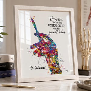 May include: Framed art print featuring a colorful watercolor illustration of a hand holding a scalpel. The artwork includes the text "Vergessen Sie nie den UNTERSCHIED, den Sie gemacht haben" and "Dr. Johnson". The print is displayed on a desk with medical books.