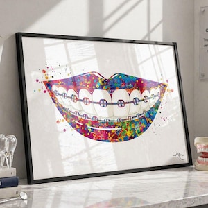 Puede incluir: Una impresión en acuarela enmarcada de una boca sonriente con brackets. La obra de arte presenta un diseño vibrante y colorido con salpicaduras de pintura. La impresión se muestra sobre un fondo blanco, con libros de temática dental y un modelo de dientes en primer plano.