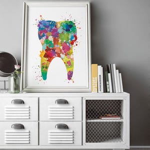 May include: A framed print featuring a colorful watercolor illustration of a tooth. The tooth is filled with vibrant splashes of red, yellow, green, blue, and purple. The artwork is displayed on a white cabinet with books and a decorative mirror.