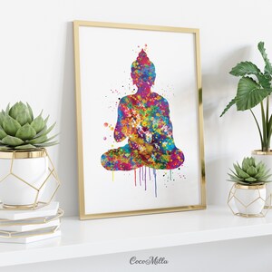 May include: A framed watercolor print of a seated Buddha figure. The artwork features a vibrant, multicolored splatter effect, with hues of blue, yellow, pink, and purple. The frame is gold, and the print is displayed on a white shelf with decorative plants.