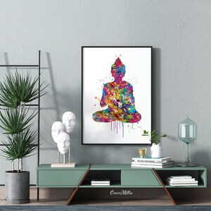 May include: A framed watercolor print of a seated Buddha in vibrant colors. The artwork features a splash of rainbow hues, with a black frame. The print is displayed on a teal-colored cabinet, with decorative items and a tall plant.