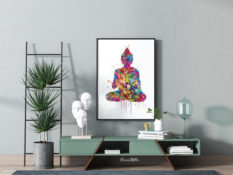 May include: A framed watercolor print of a seated Buddha in vibrant colors. The artwork features a splash of rainbow hues, with a black frame. The print is displayed on a teal-colored cabinet, with decorative items and a tall plant.