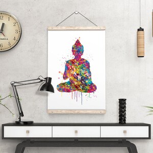 May include: A colorful watercolor print of a seated Buddha figure. The artwork features a vibrant mix of colors, including blues, greens, yellows, and reds, creating a striking visual effect. The print is displayed on a white background and framed with wooden bars.