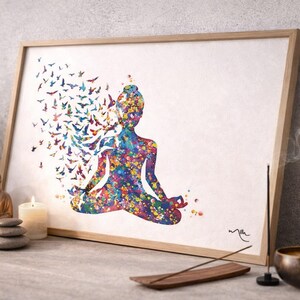 May include: A framed print depicting a person in a yoga pose, rendered in a vibrant watercolor style. The figure is made up of colorful paint splatters, with birds emanating from the head. The artwork is displayed on a table with a candle, incense, and a small Buddha statue.