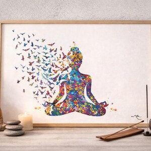 May include: A framed art print shows a colourful watercolour silhouette of a person in a yoga pose, with birds emerging from the head. The artwork features a vibrant palette of blues, purples, and yellows. The print is displayed on a table with a Buddha statue and a lit candle.