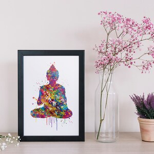 May include: A framed print featuring a colorful watercolor silhouette of a seated Buddha. The artwork is a vibrant mix of blues, greens, yellows, and reds. The print is displayed on a wooden surface, alongside a vase of pink flowers and a small potted plant.