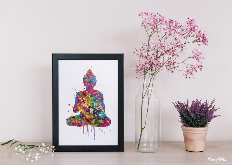 May include: A framed print featuring a colorful watercolor silhouette of a seated Buddha. The artwork is a vibrant mix of blues, greens, yellows, and reds. The print is displayed on a wooden surface, alongside a vase of pink flowers and a small potted plant.