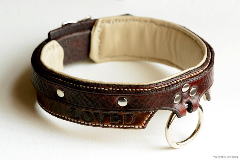 May include: A dark brown leather collar featuring a cream-colored interior lining, silver-toned metal rivets, and a central silver-toned O-ring. The word LOVED is embossed on the outer surface of the leather band, which is stitched with white thread.