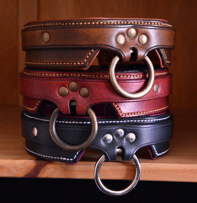 May include: A stack of three leather collars in brown, red, and black. Each collar features a wide band, decorative stitching, three metal rivets, and a large circular metal ring attached to a reinforced tab at the front center of the band.
