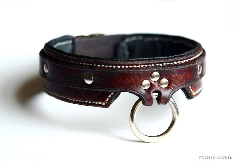 May include: A dark brown leather collar featuring white contrast stitching, silver-toned metal rivets, and a central silver-toned O-ring. The text TWISTED LEATHER is printed in the bottom right corner of the image.