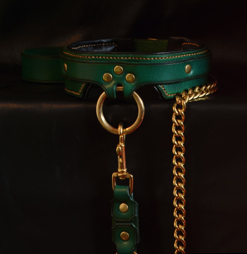 May include: A dark green leather collar featuring gold-toned metal hardware, including a central O-ring, rivets, and a detachable gold-toned chain leash. The collar has visible orange stitching along the edges and a black interior lining.