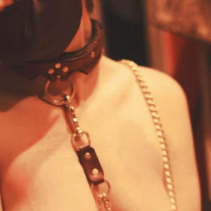 May include: A black leather collar featuring silver-toned metal hardware, including a central O-ring and a vertical strap attachment. A silver-toned metal chain is connected to the collar, extending downwards and across the chest area.