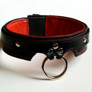 May include: A black leather collar featuring a vibrant red interior lining and red stitching along the edges. The collar includes silver-toned metal rivets and a prominent silver-toned metal O-ring attached to the front. The text TWISTED LEATHER is visible.