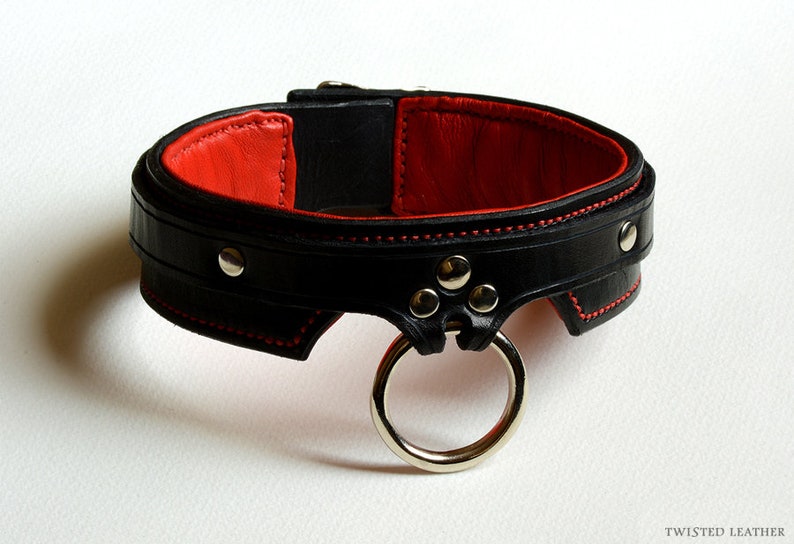 May include: A black leather collar featuring a vibrant red interior lining and red stitching along the edges. The collar includes silver-toned metal rivets and a prominent silver-toned metal O-ring attached to the front. The text TWISTED LEATHER is visible.