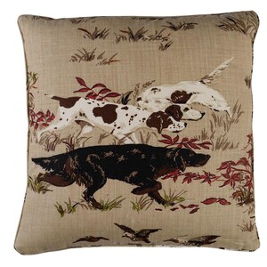 May include: Beige throw pillow with a hunting scene featuring three dogs running through a field of brown and green grass. The dogs are a black Labrador Retriever, a brown and white pointer, and a white pointer. The background is a beige linen fabric.