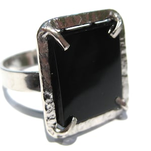 Puede incluir: Un anillo de tono plateado con una piedra negra rectangular grande engastada en un marco de metal martillado. La piedra se sujeta con cuatro garras en las esquinas, y la banda del anillo es lisa y pulida, contrastando con el engaste texturizado.