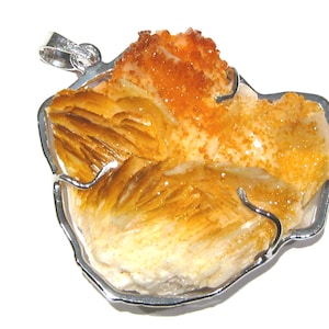 Puede incluir: Un colgante de plata con un cristal natural naranja y amarillo de forma irregular y rugosa. El cristal está engastado en un marco de plata y tiene un bucle en la parte superior para sujetar una cadena.