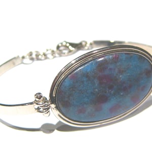 Puede incluir: Una pulsera de plata con una gran piedra ovalada azul y morada engastada en un bisel de plata. La pulsera tiene un cierre de cadena.