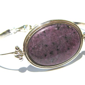 Puede incluir: Un brazalete de plata con una gran piedra ovalada de color púrpura engastada en un bisel de plata. La piedra preciosa tiene un patrón moteado.