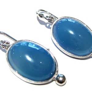 May include: A pair of oval earrings featuring a light blue, glossy stone set in a silver-coloured metal frame. The earrings have a hook closure and a small, round accent at the bottom. The jewellery is displayed against a white background.