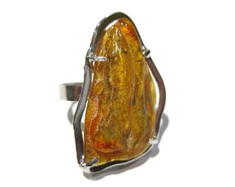 May include: A silver ring featuring a large, teardrop-shaped amber stone. The amber is a warm, translucent yellow-orange color with natural inclusions. The ring has a simple, modern design.