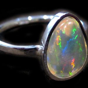 Op de afbeelding: Een zilveren ring met een peervormige opaal edelsteen. De opaal toont een levendig kleurenspel, met groene, blauwe en rode flitsen. De ring heeft een gladde, gepolijste band, tegen een donkere achtergrond.