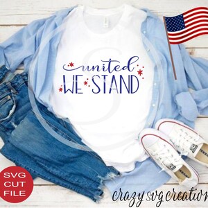 May include: A white t-shirt with the text "united we stand" in blue with red stars. The shirt is laid out on a wooden surface with blue jeans, a light blue shirt, and white sneakers.