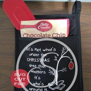 K&ouml;nnte beinhalten: Schwarzer Ofenhandschuh mit einer Tasche, die einen roten Spatel und eine Betty Crocker Chocolate Chip-Keksmischung enth&auml;lt. Der Handschuh hat eine wei&szlig;e Grafik mit einem Weihnachtszitat und einem Charlie Brown-Bezug.
