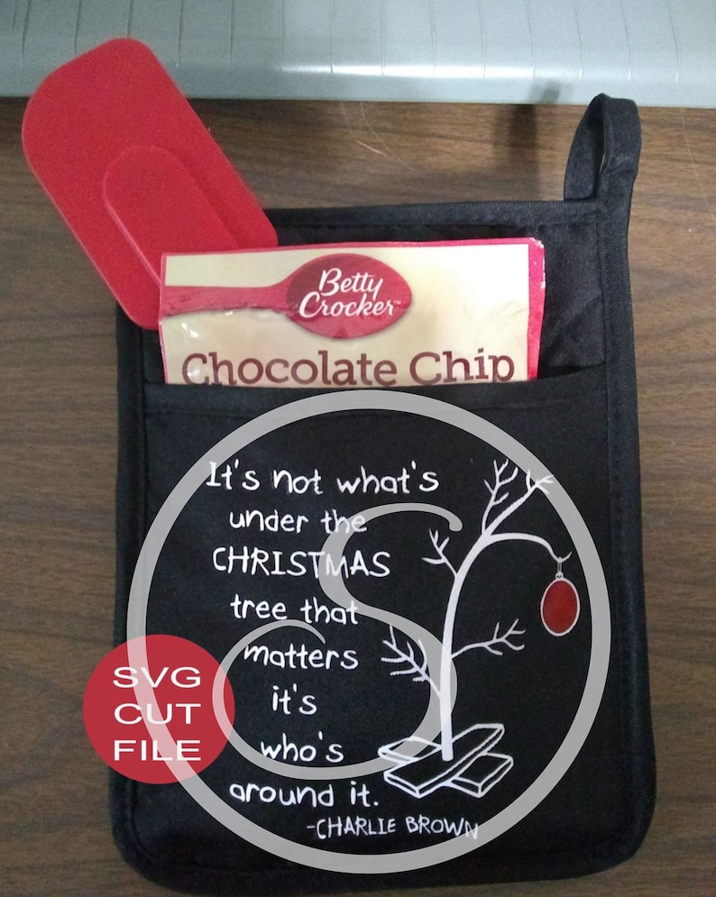 K&ouml;nnte beinhalten: Schwarzer Ofenhandschuh mit einer Tasche, die einen roten Spatel und eine Betty Crocker Chocolate Chip-Keksmischung enth&auml;lt. Der Handschuh hat eine wei&szlig;e Grafik mit einem Weihnachtszitat und einem Charlie Brown-Bezug.