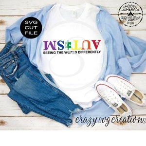 Puede incluir: Una camiseta blanca con el texto "WS#LOV" en colores arco&iacute;ris y la frase "SEEING THE WORLD DIFFERENTLY" en texto negro. La camiseta est&aacute; colocada sobre una superficie de madera con unos vaqueros azules, una camisa azul claro y unas zapatillas blancas.