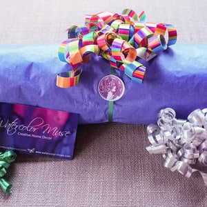 May include: A blue gift wrapped in tissue paper with a colorful ribbon and a sticker that says "Watercolor Muse Creative Home Decor" and the website www.watercolor-muse.com. The gift is surrounded by green, silver, and purple curling ribbon.
