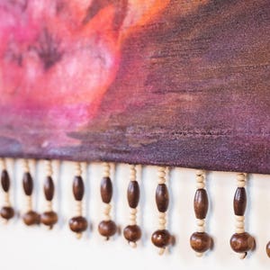 May include: A close-up of a purple fabric with a pink and orange watercolor design. The fabric has a fringe of brown wooden beads.
