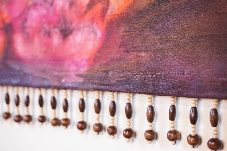 May include: A close-up of a purple fabric with a pink and orange watercolor design. The fabric has a fringe of brown wooden beads.