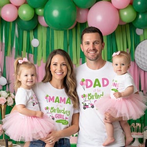 May include: Family of four wearing matching white t-shirts with golf-themed birthday graphics. The shirts read "MAMA of the Birthday Girl" and "DAD of the Birthday". The two girls wear pink tutus. The background includes balloons and golf course decorations.