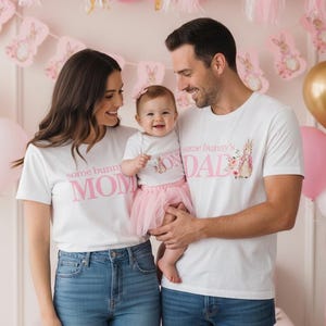 May include: A family of three wearing matching white t-shirts and blue jeans. The shirts have pink text that reads "some bunny's MOM", "some bunny's DAD", and "some bunny". The baby wears a pink tutu. Pink and gold balloons are in the background.