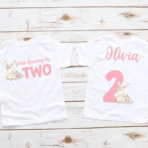 May include: Two white toddler t-shirts with pink text and bunny graphics. One shirt says "Some bunny is TWO" with a bunny and floral design. The other shirt has the name "Olivia" and a large pink number "2" with a bunny.