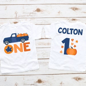 May include: Two white t-shirts with a fall theme. The front of one shirt has a blue truck with orange pumpkins and the text "ONE". The back of the other shirt has the name "COLTON" and the number "1" with a pumpkin and orange leaves.