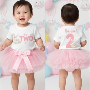 May include: A baby wearing a white t-shirt with the text "hop TWO it!" and a pink tutu. The shirt has a bunny and floral design. The back of the shirt has the name "Hannah" and the number "2". Balloons and bunny decorations are in the background.