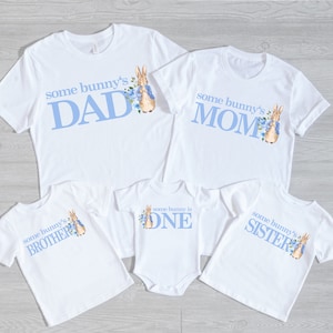 Puede incluir: Conjunto de cinco camisetas blancas con texto azul y una ilustraci&oacute;n de Peter Rabbit. Las camisetas dicen "some bunny's DAD", "some bunny's MOM", "some bunny's BROTHER", "some bunny is ONE", y "some bunny's SISTER".