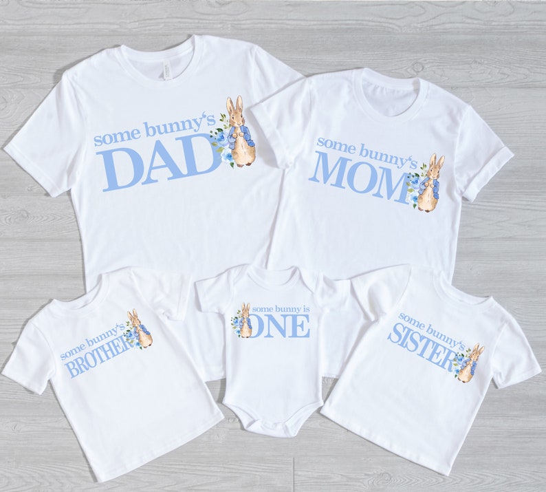 Puede incluir: Conjunto de cinco camisetas blancas con texto azul y una ilustraci&oacute;n de Peter Rabbit. Las camisetas dicen "some bunny's DAD", "some bunny's MOM", "some bunny's BROTHER", "some bunny is ONE", y "some bunny's SISTER".
